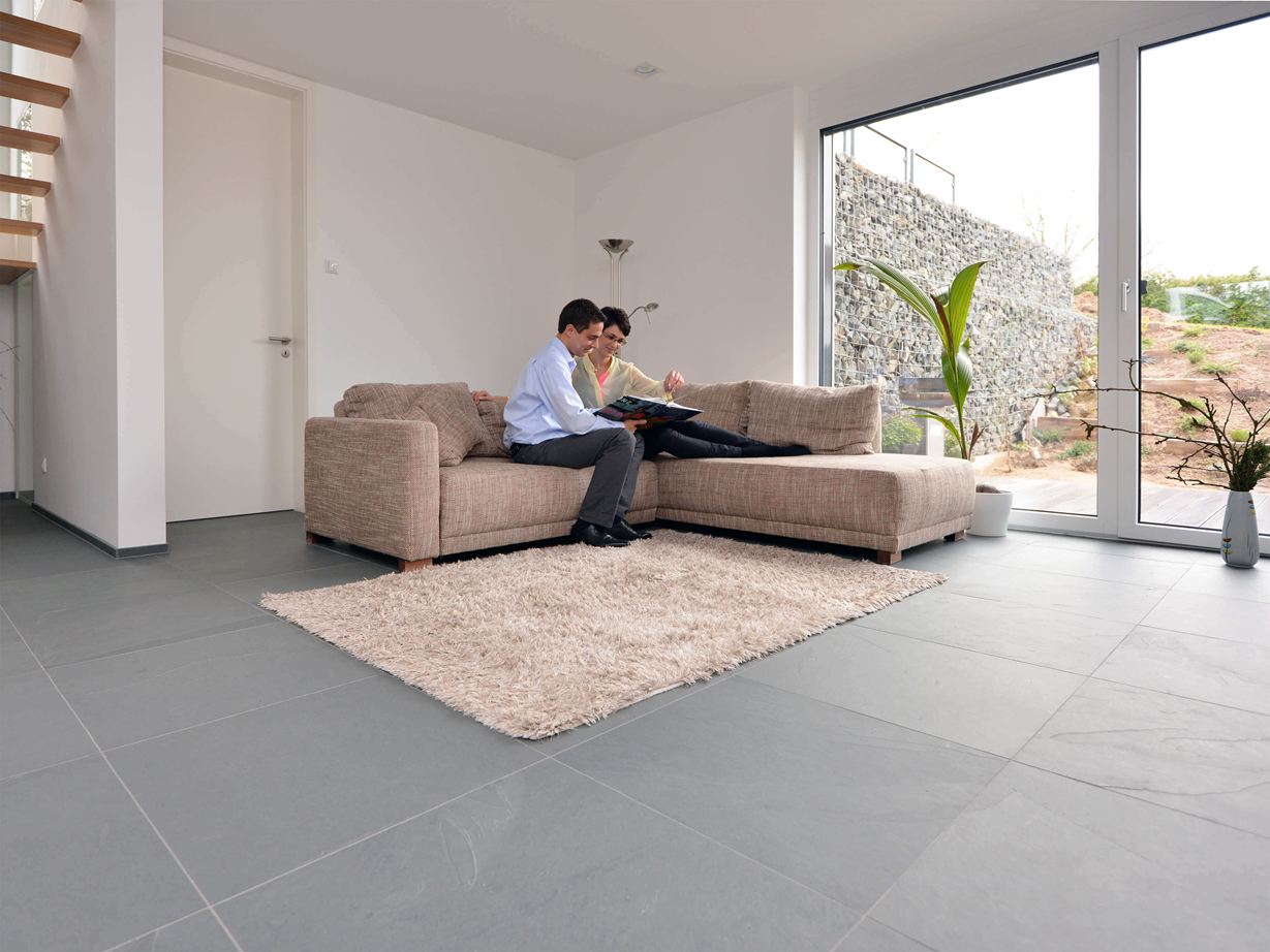 La modernité du sol est accentuée par les joints fins Ardoise grand format Grey Slate dans le salon avec canapé et murs blancs. Un couple est assis sur le canapé