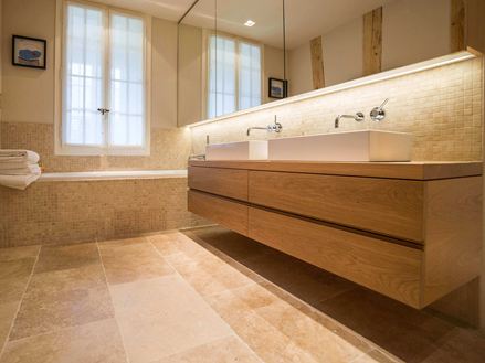 Salle de bain moderne avec lavabo en bois, grandes fenêtres et carreaux de travertin beige.
