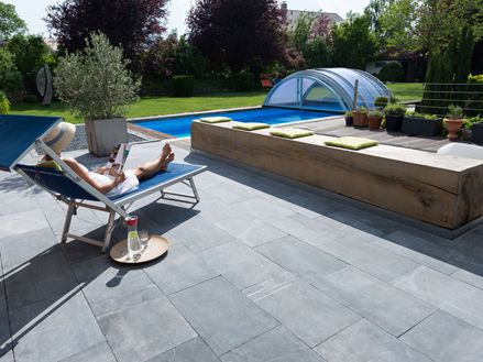 Femme se détendant sur une chaise longue au bord de la piscine, dalles de terrasse en pierre bleue et jardin en arrière-plan.